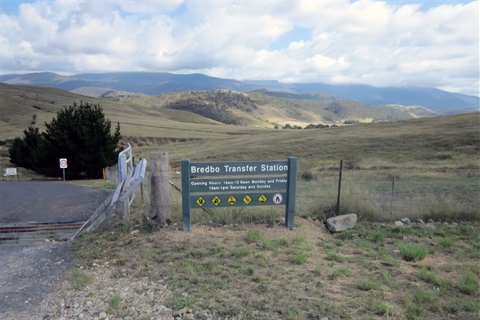 Wooden Bredbo Transfer Station sign with rolling hills and paddocks beneath a cloudy blue sky