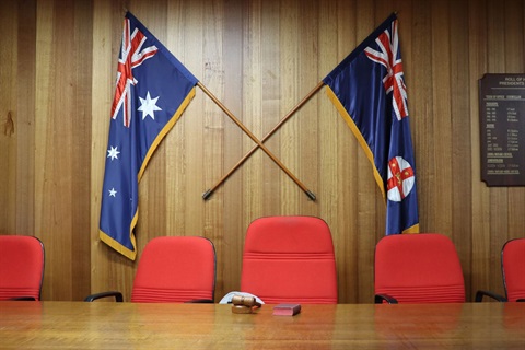 Cooma Council Chambers, head of the meeting table with empty chairs and councillor paraphernalia and state/national flags behind.