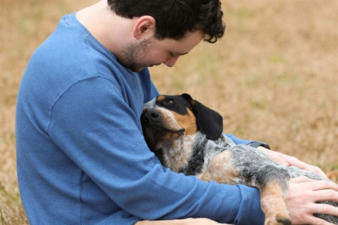 A man with short curly dark brown hair cuddling a large dog outdoors