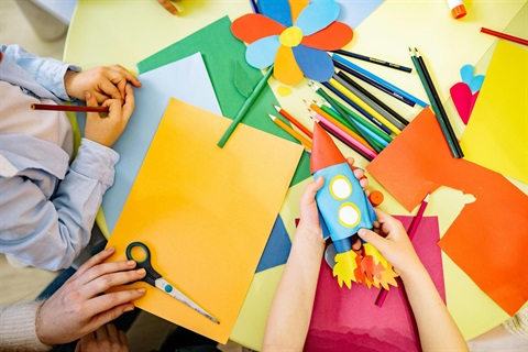 Flatlay image of preschoolers making papercrafts on a table covered in colourful paper