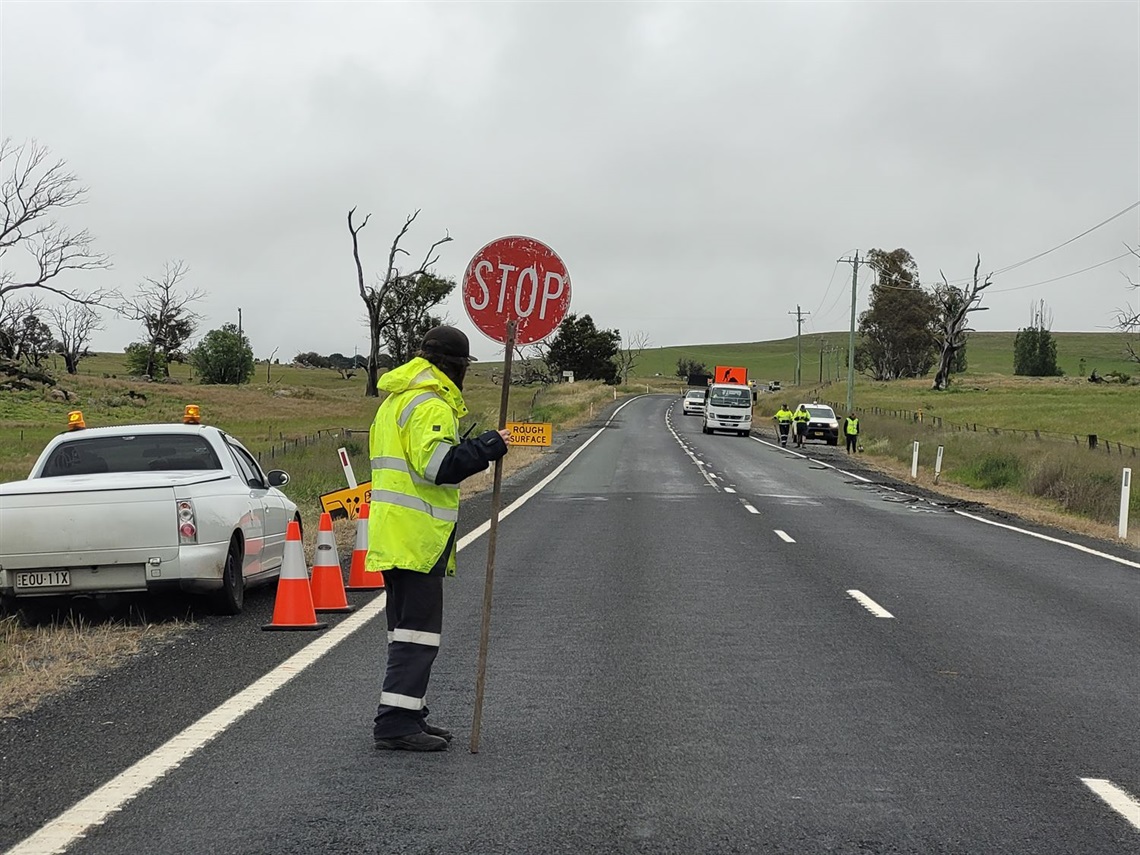 A traffic controller in a high visibility raincoat holds a stop sign standing on a two-lane road on an overcast day, a work truck is in the background and a ute to his left