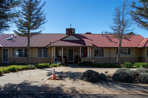 The front exterior of Snowy River Hostel in Berridale, a single-level brick-veneer building with quaint gardens and eucalypt trees.