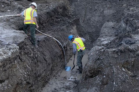 Two workers wearing high-vis gear and hard-hats repair a rural water main in a deep trench